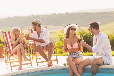Téléchargez les photos : Groupe d'amis à une fête d'été au bord de la piscine, assis au bord d'une piscine, boire des cocktails et de la bière et s'amuser - en image libre de droit