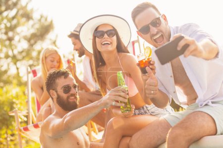 Téléchargez les photos : Groupe d'amis s'amuser à une fête d'été au bord de la piscine, assis au bord d'une piscine, boire des cocktails et de la bière et prendre des selfies - en image libre de droit