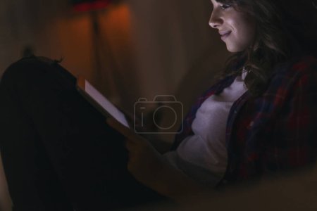 Beatiful young woman at home, sitting in the dark on a living room sofa, surfing the net on a tablet computer, enjoying her free time