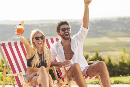 Téléchargez les photos : Couple amoureux à une fête d'été au bord de la piscine, assis sur des chaises longues au bord de la piscine, boire des cocktails et saluer les amis qui arrivent à la fête - en image libre de droit