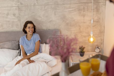 Téléchargez les photos : Mari surprenant sa femme en apportant son petit déjeuner et ses fleurs au lit sur un plateau ; couple relaxant à la maison le matin - en image libre de droit