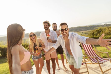 Foto de Grupo de jóvenes amigos divirtiéndose en una fiesta de verano junto a la piscina, bebiendo cerveza e invitando a más amigos a unirse a ellos - Imagen libre de derechos