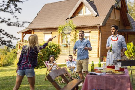Photo for Group of friends having a backyard barbecue party, grilling meat, drinking beer, playing badminton and having fun on a sunny summer day. Focus on the men - Royalty Free Image