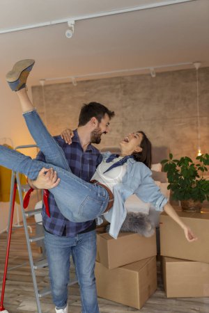 Photo for Beautiful young couple in love moving in together, having fun while unpacking cardboard boxes with their belongings, boyfriend carrying girlfriend in his arms - Royalty Free Image