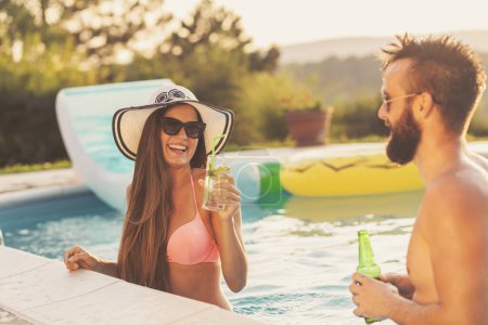 Foto de Pareja enamorada en una fiesta de verano junto a la piscina, parada en el agua junto al borde de la piscina, bebiendo cerveza y divirtiéndose - Imagen libre de derechos