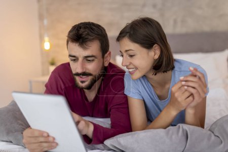 Photo for Beautiful couple lying in bed and surfing the Net on a tablet computer, searching for home decor ideas - Royalty Free Image