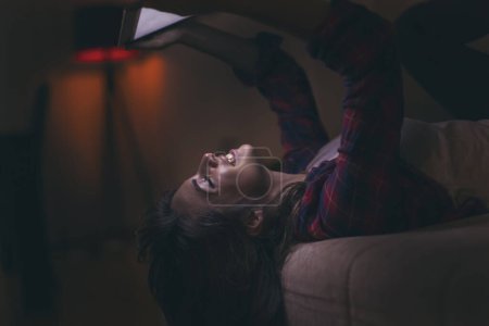 Foto de Mujer joven acostada en un sofá de la sala de estar por la noche, navegando por la web en una tableta, disfrutando de su tiempo libre - Imagen libre de derechos