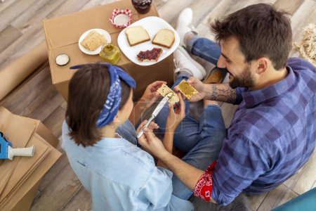 Photo for Top view of young couple in love sitting on the floor among cardboard boxes, having fun while eating breakfast in their new apartment - Royalty Free Image