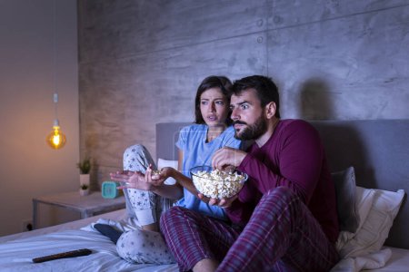 Téléchargez les photos : Beau jeune couple amoureux, assis au lit côte à côte, mangeant du pop-corn et s'amusant à regarder un film d'horreur la nuit - en image libre de droit