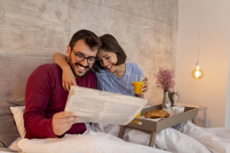 Foto de Pareja enamorada vistiendo pijamas, sentada en la cama por la mañana, leyendo periódicos y desayunando, relajándose en casa - Imagen libre de derechos