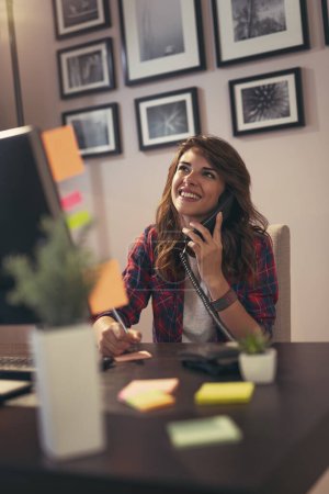 Téléchargez les photos : Jeune femme travaillant dans un bureau à domicile, ayant une conversation téléphonique - en image libre de droit