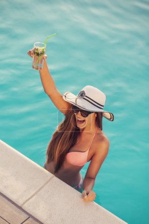 Téléchargez les photos : Jolie jeune femme portant un bikini, des lunettes de soleil et un chapeau, debout dans la piscine à côté du bord, boire des cocktails et danser - en image libre de droit