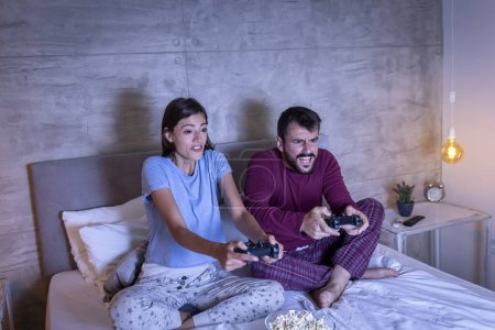 Foto de Hermosa pareja joven usando pijamas, divirtiéndose en casa, jugando videojuegos y comiendo palomitas de maíz en la cama por la noche - Imagen libre de derechos
