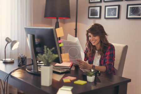 Foto de Mujer joven sentada en un escritorio en su oficina en casa, sosteniendo documentos alegres después de un éxito empresarial - Imagen libre de derechos
