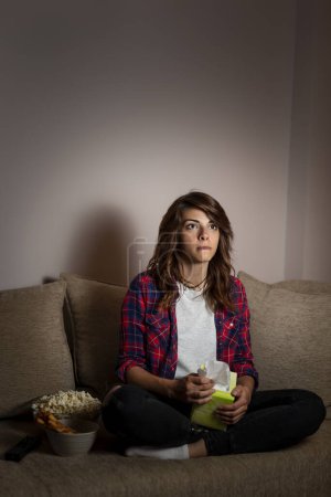 Téléchargez les photos : Belle jeune femme assise dans le noir sur un canapé du salon, regardant un film dramatique - en image libre de droit