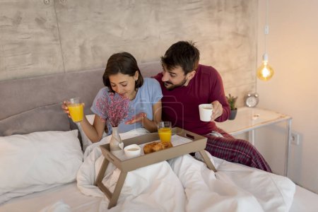 Téléchargez les photos : Beau jeune couple amoureux en pyjama assis au lit, homme femme surprenante en apportant son petit déjeuner et des fleurs au lit sur un plateau - en image libre de droit
