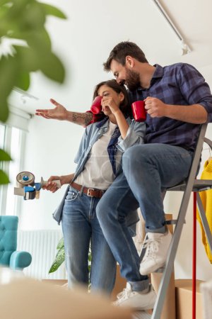 Photo for Beautiful young couple in love drinking coffee while moving in together into new apartment, sitting among unpacked boxes, planning apartment redecoration - Royalty Free Image