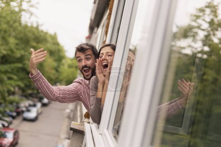Téléchargez les photos : Beau jeune couple penché par une fenêtre ouverte de l'appartement, saluant et criant, appelant des amis passant dans la rue - en image libre de droit
