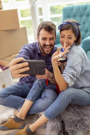 Téléchargez les photos : Jeune couple amoureux déménageant dans un nouvel appartement, buvant du vin et s'amusant à prendre des selfies avec les clés de l'appartement - en image libre de droit