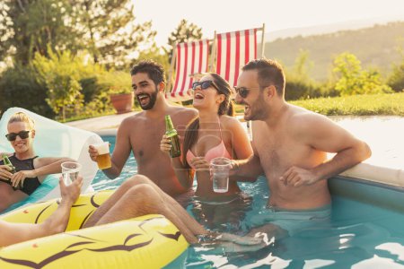 Group of friends at a poolside summer party, having fun in the swimming pool, drinking cocktails and beer and making a toast