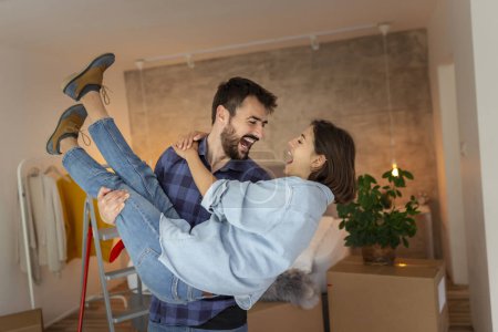 Photo for Beautiful newly married young couple moving in together, having fun while unpacking cardboard boxes with their belongings, husband carrying wife in his arms - Royalty Free Image