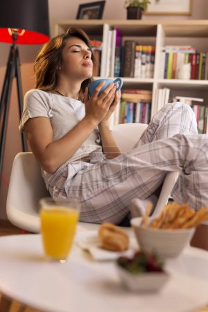 Foto de Hermosa joven con pijama, sentada junto a la ventana de la sala de estar, sosteniendo una taza de café y disfrutando del momento - Imagen libre de derechos