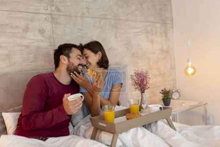 Photo for Beautiful young couple in love wearing pajamas sitting in bed, hugging and kissing while drinking coffee, having breakfast and relaxing at home in the morning - Royalty Free Image