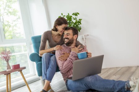 Téléchargez les photos : Couple amoureux se détendre à la maison, câlins, boire du café et surfer sur le net sur un ordinateur portable - en image libre de droit
