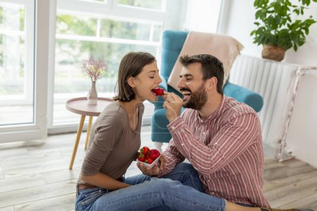Téléchargez les photos : Beau jeune couple amoureux assis par terre dans leur nouvelle maison, mangeant des fraises, riant et s'amusant - en image libre de droit