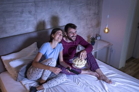 Téléchargez les photos : Couple amoureux couché au lit, manger du pop-corn et regarder un film, se détendre à la maison la nuit - en image libre de droit