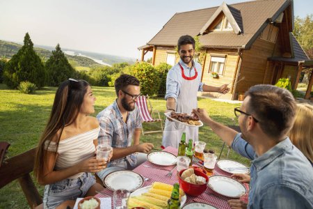 Photo for Group of friends having an outdoor barbecue lunch, eating grilled meat, drinking beer and having fun. Focus on the man standing - Royalty Free Image