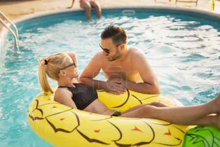 Téléchargez les photos : Couple amoureux à une fête d'été au bord de la piscine, bronzer et s'amuser ; fille flottant dans la piscine sur un matelas pneumatique en forme d'ananas - en image libre de droit