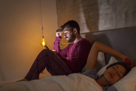 Foto de Hombre usando un teléfono inteligente mientras su novia duerme en la cama por la noche; insomnio y concepto de engaño - Imagen libre de derechos