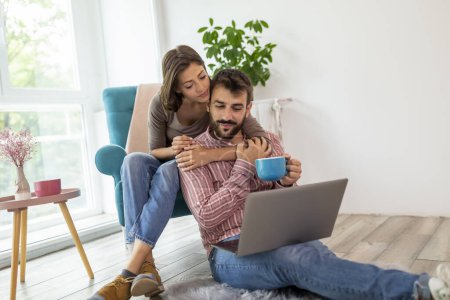 Téléchargez les photos : Couple amoureux assis sur le sol, boire du café et à la recherche d'idées de décoration à la maison sur un ordinateur portable - en image libre de droit