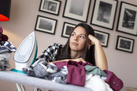 Young bored and tired woman leaning on the ironing board, surrounded with bunch of wrinkled clothes ready for ironing