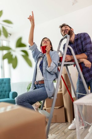 Photo for Couple in love moving in together, woman sitting on the ladder, drinking coffee and pointing to the place on the ceiling for lighting arrangement - Royalty Free Image