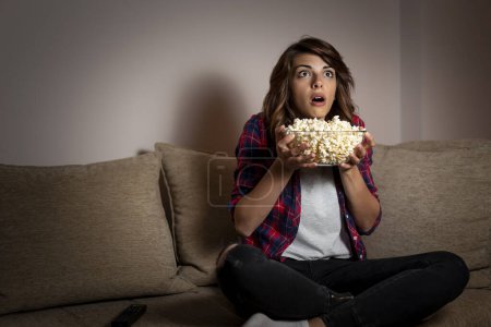 Téléchargez les photos : Belle jeune femme assise dans le noir sur un canapé du salon, s'amusant à regarder la télévision et à manger du pop-corn - en image libre de droit