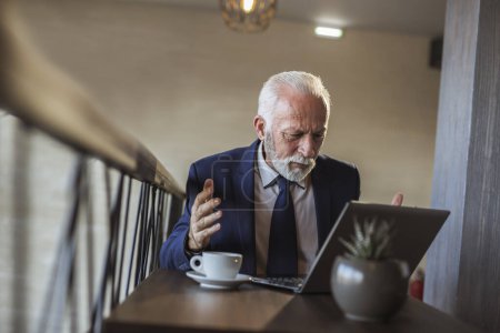 Photo for Senior businessman in a restaurant, analysing financial data and reports using laptop computer, displeased and angry - Royalty Free Image