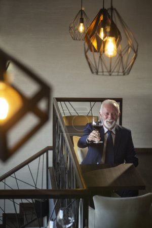 Téléchargez les photos : Homme d'affaires senior assis à une table de restaurant, lever un verre de vin et porter un toast après avoir fait une affaire réussie - en image libre de droit