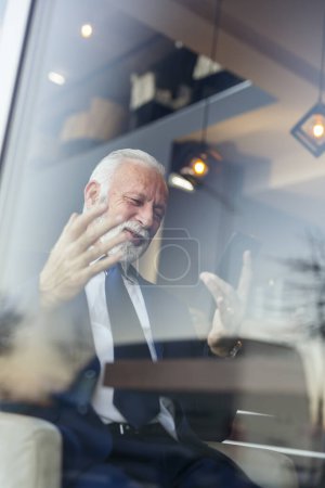Téléchargez les photos : Homme d'affaires senior assis à une table de restaurant, en utilisant un téléphone intelligent, satisfait après avoir reçu de bonnes nouvelles - en image libre de droit
