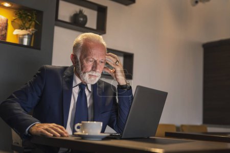 Foto de Hombre de negocios sénior sentado en una mesa de restaurante, trabajando en una computadora portátil y tomando una taza de café - Imagen libre de derechos