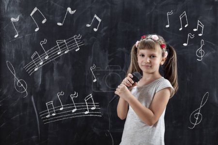 Téléchargez les photos : Écolière debout devant un tableau noir, tenant un micro et chantant sur une leçon de musique - en image libre de droit
