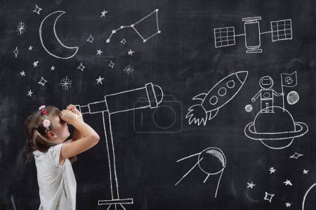 Photo for Schoolgirl standing in front of a blackboard, watching stars through a chalk-drawn telescope, learning about space and astronomy - Royalty Free Image