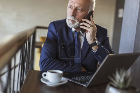 Foto de Hombre de negocios senior que trabaja en una oficina moderna, hablando por teléfono mientras trabaja en una computadora portátil - Imagen libre de derechos