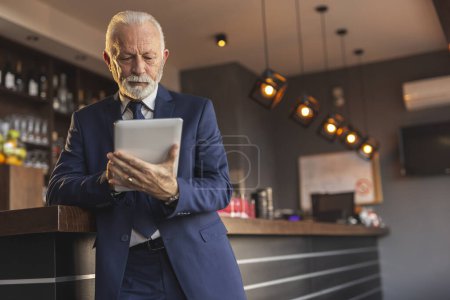 Photo for Senior restaurant manager standing next to a counter, using a tablet computer - Royalty Free Image