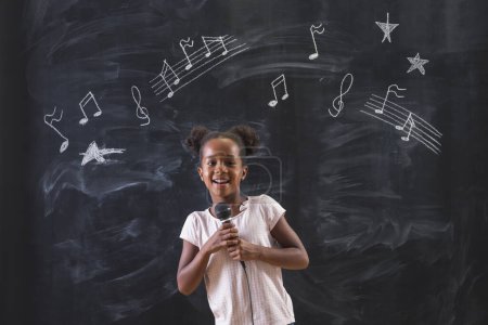 Téléchargez les photos : Belle écolière mixte du primaire debout devant un tableau en classe, chantant en classe de musique avec des symboles de notes musicales dessinés sur le tableau noir - en image libre de droit