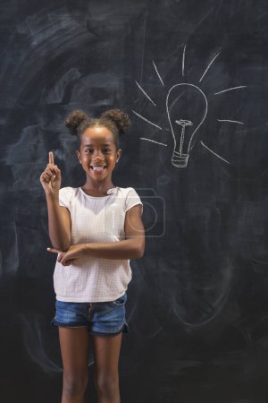 Photo for Elementary school girl standing in front of a chalkboard in classroom, happy after coming up with an idea, holding index finger up with light bulb drawn on the blackboard behind her - Royalty Free Image