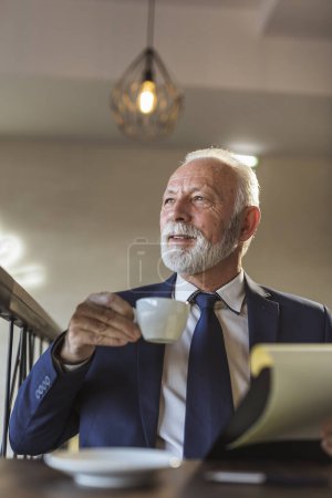Photo for Senior businessman sitting in a modern office building restaurant, reading new contract terms and conditions and drinking coffee - Royalty Free Image