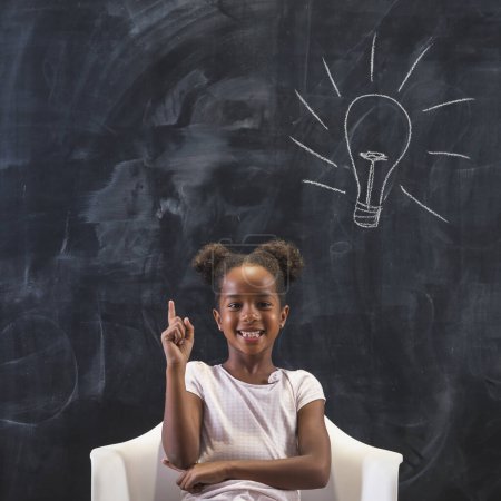 Photo for Elementary school girl sitting in front of a chalkboard in classroom, happy after coming up with an idea, holding index finger up with light bulb drawn on the blackboard behind her - Royalty Free Image
