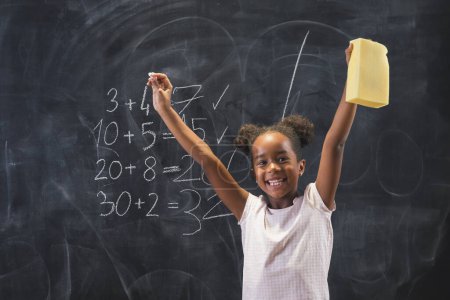 Beautiful mixed race elementary school girl standing in front of a chalkboard, happy after solving math equations and getting an A, smiling with hands up and fists clenched